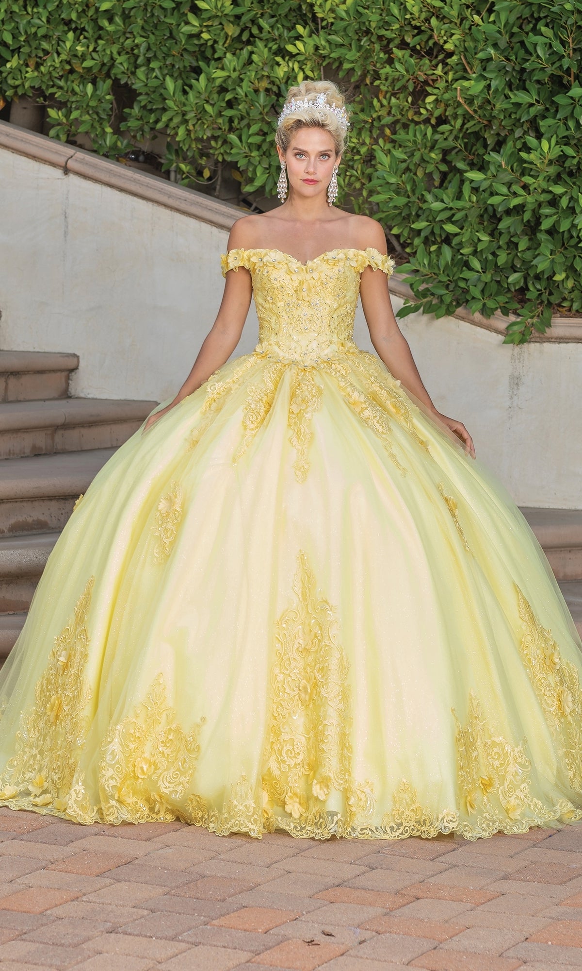 A woman wears the Quinceanera Dress 1739 By Dancing Queen, a yellow off-the-shoulder gown with gold embellishments and lace embroidery, standing on a stone path before stairs and greenery, accessorized with a tiara and large earrings.